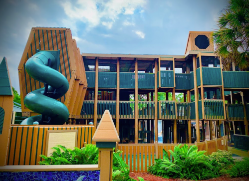 Colorful playground structure with a green slide, surrounded by palm trees and lush greenery under a cloudy sky.