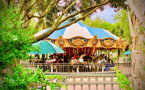 A colorful carousel surrounded by greenery and umbrellas, with people enjoying a sunny day at the park.