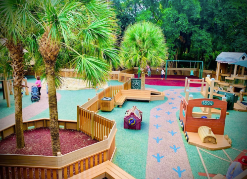 A colorful playground with palm trees, wooden structures, and children playing in a lush green environment.