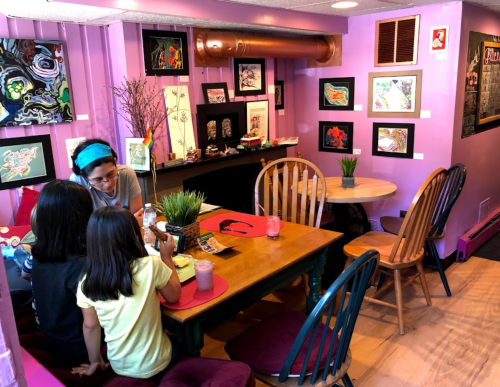 A cozy café with purple walls, featuring artwork, a wooden table, and two children chatting with an adult.