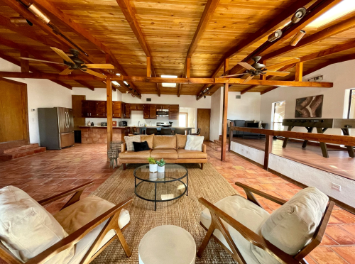 Spacious living area with wooden beams, a brown sofa, two chairs, and a kitchen in the background. Warm, inviting atmosphere.