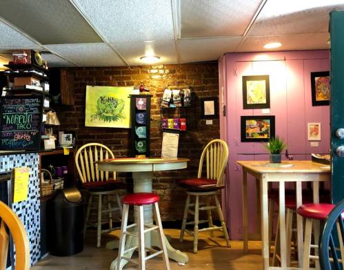 Cozy café interior with wooden tables, colorful artwork, and a brick wall, featuring a purple cabinet and plants.