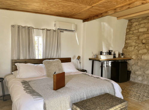 Cozy bedroom with a large bed, wooden accents, and a stone wall, featuring a small kitchenette and natural light.