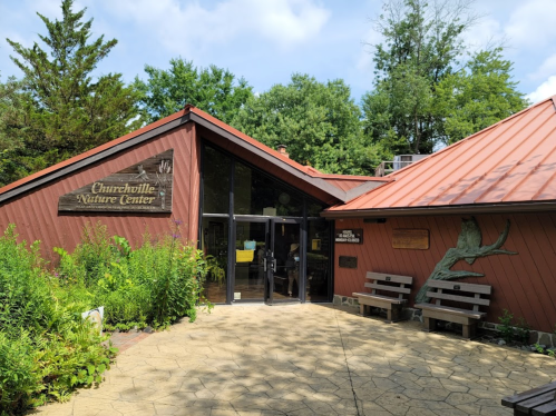 Exterior of Churchillville Nature Center with a sloped roof, wooden signage, and benches in a green, landscaped area.