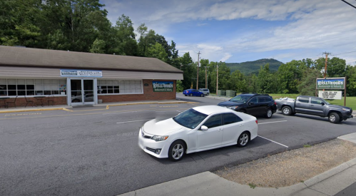 A white car parked in front of a restaurant with a sign, surrounded by trees and mountains in the background.