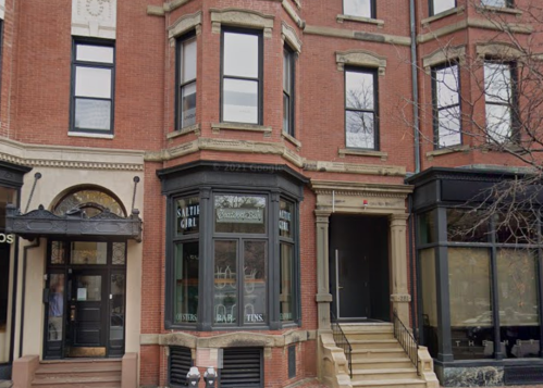 A brick building with large windows, featuring a staircase entrance and storefronts on either side.