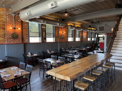 Interior of a restaurant featuring wooden tables, black chairs, exposed brick walls, and a staircase.