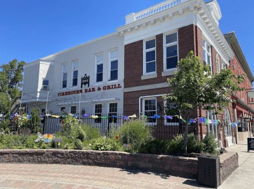 A brick building with "Firehouse Bar & Grill" sign, surrounded by greenery and colorful decorations on a sunny day.