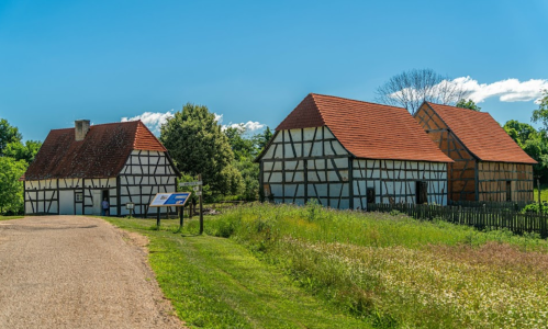 Two traditional timber-framed houses with red roofs beside a gravel path, surrounded by green grass and trees.