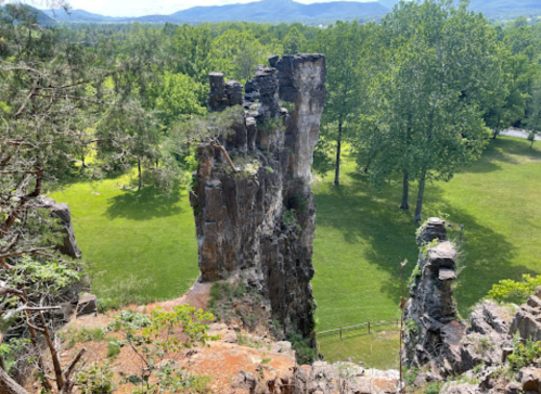 A rocky cliff rises above a green park, surrounded by trees and mountains in the background on a sunny day.