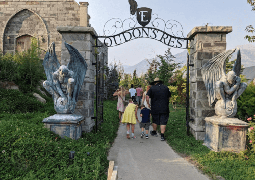 A group of children and adults walk through a stone archway with winged statues, entering a whimsical garden.