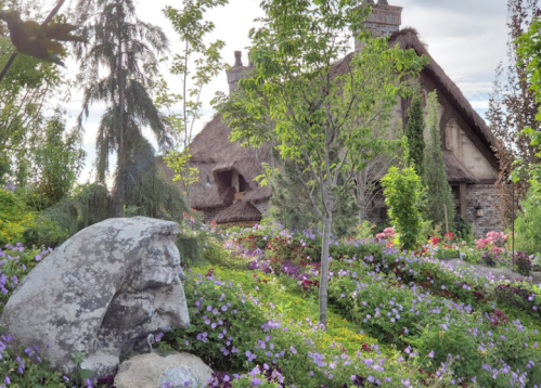 A picturesque garden with colorful flowers, a stone sculpture, and a charming thatched-roof house in the background.