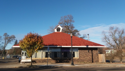 A round brick building with a red roof and a small steeple, surrounded by trees under a clear blue sky.