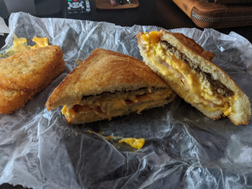 A toasted sandwich filled with melted cheese, ham, and egg, alongside two golden hash browns on crinkled paper.