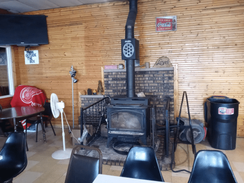 Cozy interior of a restaurant featuring a brick fireplace, wooden walls, and simple seating arrangements.