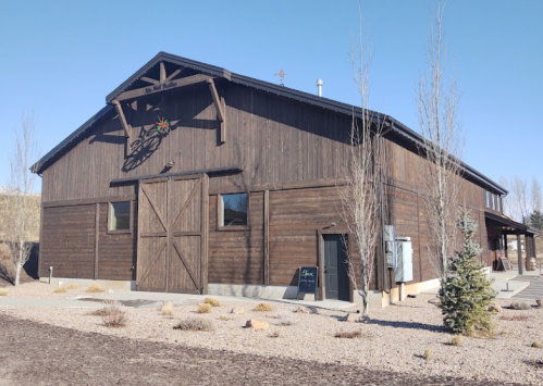 A large wooden barn with a decorative star, surrounded by gravel and sparse landscaping under a clear blue sky.