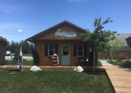 A wooden cabin with a sign reading "simplyeden," surrounded by greenery and a clear blue sky.