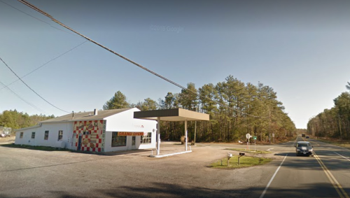 A small, colorful building with a gas station canopy beside a rural road, surrounded by trees.