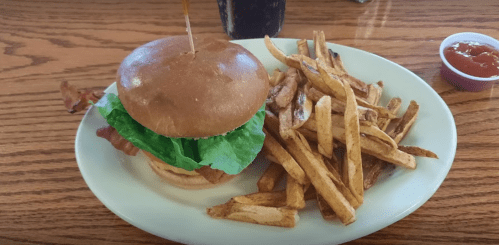 A plate with a burger topped with lettuce and bacon, served with a side of golden fries and a small cup of ketchup.