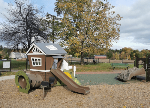 Playground with a small playhouse, slide, and climbing structure surrounded by trees and benches.