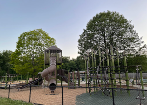 A playground featuring a large slide, climbing structures, and green trees, enclosed by a fence at dusk.
