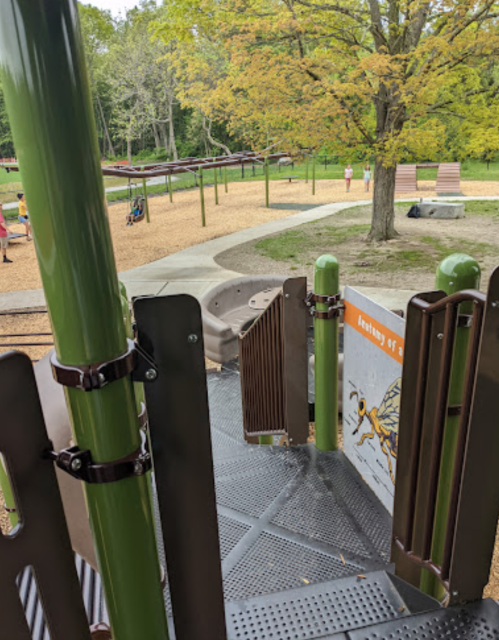 View from a playground slide, showing climbing structures and a park with trees in the background.