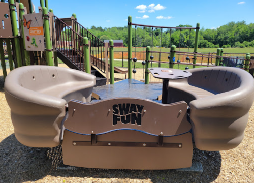 Two curved benches with a central table, labeled "SWAY FUN," in a playground setting with green trees in the background.