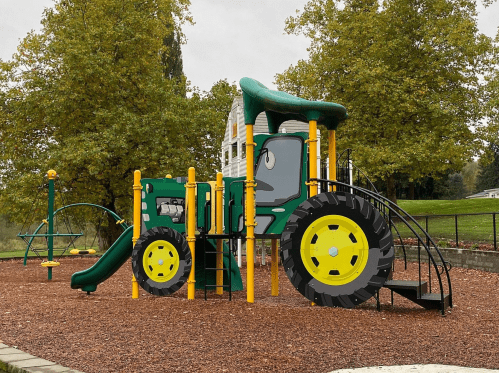 A colorful playground structure designed like a tractor, featuring slides and climbing areas, surrounded by trees.