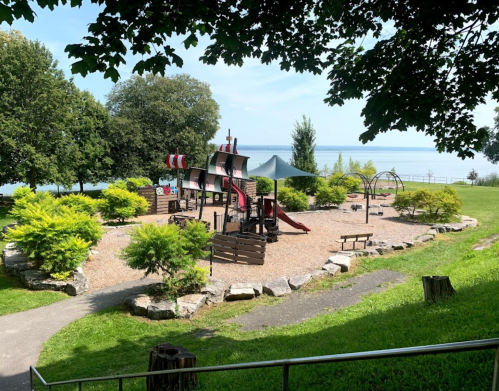 A playground with pirate-themed equipment surrounded by greenery, overlooking a calm lake on a sunny day.