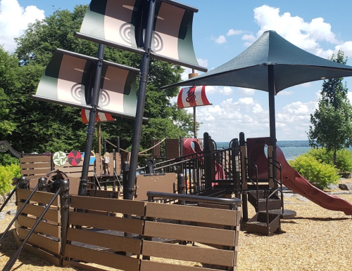 A playground with a pirate ship theme, featuring slides, climbing structures, and colorful sails against a sunny sky.