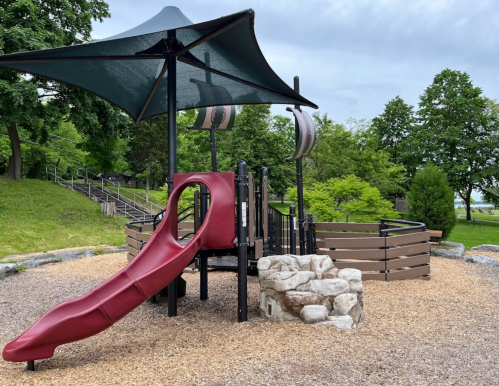 A playground featuring a red slide, climbing structure, and shaded area, surrounded by trees and gravel.