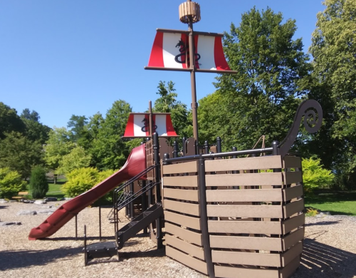 A playground structure resembling a pirate ship with a slide and sails, surrounded by greenery under a clear blue sky.