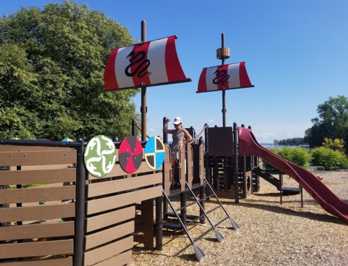 A playground with pirate ship features, including sails and a slide, set against a clear blue sky and green trees.