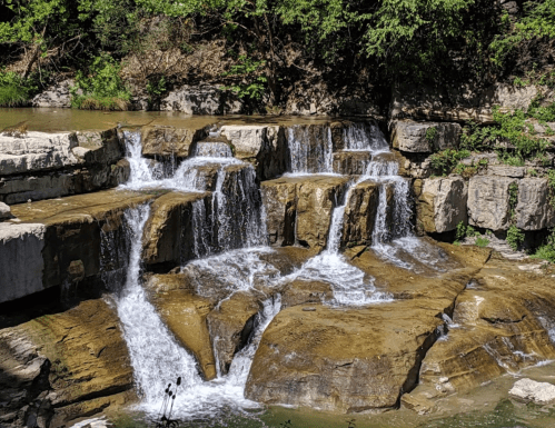 A serene waterfall cascading over rocky ledges, surrounded by lush greenery and a calm pool below.