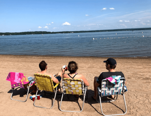 Three people sit in beach chairs on a sandy shore, enjoying drinks and looking out at a calm lake under a blue sky.