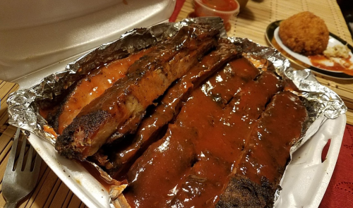 A takeout container filled with barbecue ribs smothered in sauce, alongside a side dish.
