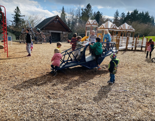 Children playing on a blue airplane structure at a playground, with various play equipment and a wooden building in the background.