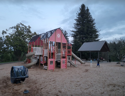 A colorful playground resembling a barn, with slides and climbing structures, set in a park with trees in the background.