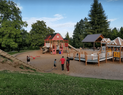 A colorful playground with slides and climbing structures, surrounded by trees and children playing.