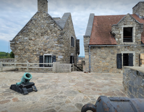 Historic stone buildings with a cannon in the foreground, set against a cloudy sky.
