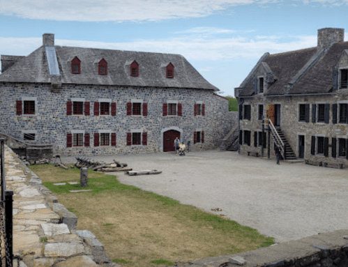Historic stone buildings surround a grassy courtyard under a cloudy sky, with visitors exploring the area.