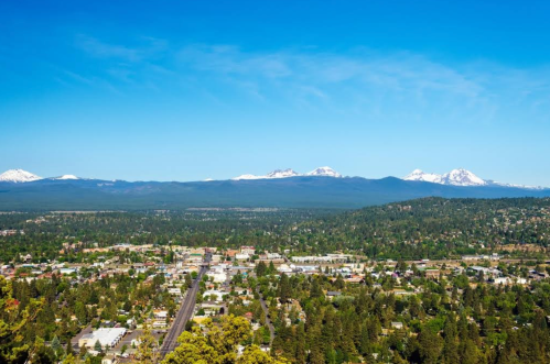 A panoramic view of a town surrounded by lush greenery and snow-capped mountains under a clear blue sky.