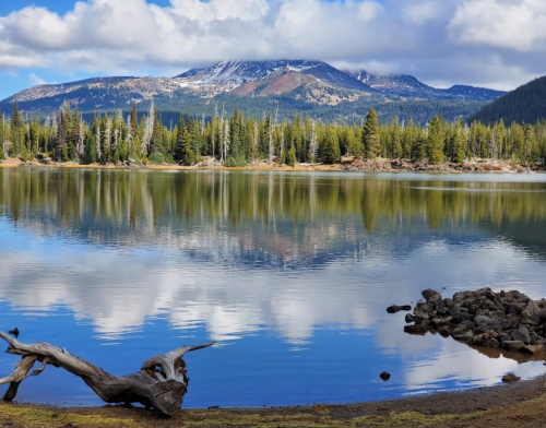 A serene lake reflects a mountain and clouds, surrounded by lush trees and rocky shoreline.