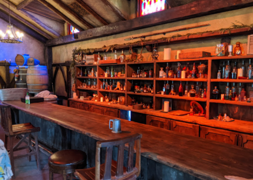 A rustic bar with wooden shelves filled with colorful bottles and jars, set in a cozy, warmly lit room.