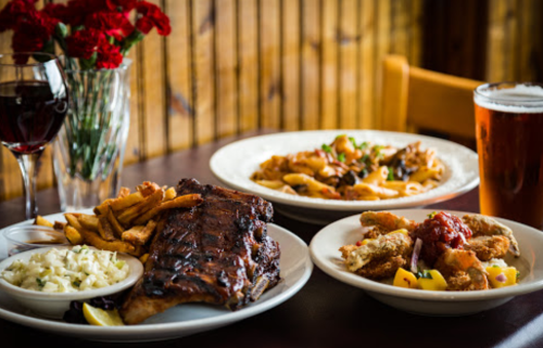 A table set with a plate of ribs, coleslaw, fries, and a side of fried food, alongside wine and beer.