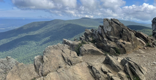 A panoramic view of green mountains under a partly cloudy sky, with rocky terrain in the foreground.