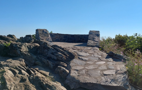 A stone observation platform on rocky terrain, surrounded by greenery under a clear blue sky.