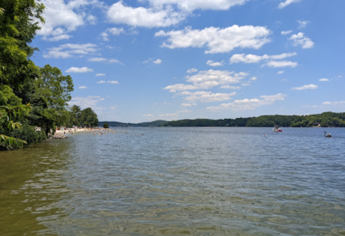 A serene lake scene with clear blue skies, fluffy clouds, and lush greenery along the shore.