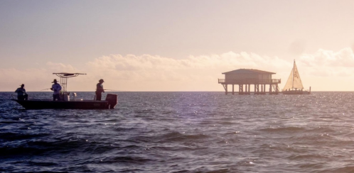 Two fishermen on a boat near a pier house, with a sailboat in the distance under a golden sunset sky.