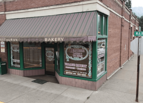 A corner bakery with a green awning and decorative signage, featuring large windows and a brick exterior.